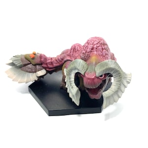 May include: A pink and white monster figurine with large horns. The figurine is posed on a black base.