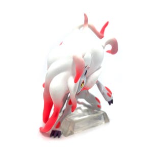 Puede incluir: Figura blanca y roja de una criatura m&iacute;tica en posici&oacute;n agachada. La figura tiene un cuerpo blanco con detalles rojos en las orejas, patas y cola. Est&aacute; colocada sobre una base rectangular transparente.