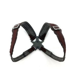 May include: A dark gray and brown harness with metal buckles and a central connector. The harness features brown straps and gray fabric, designed for a secure fit. The design is symmetrical and appears to be made of durable materials.