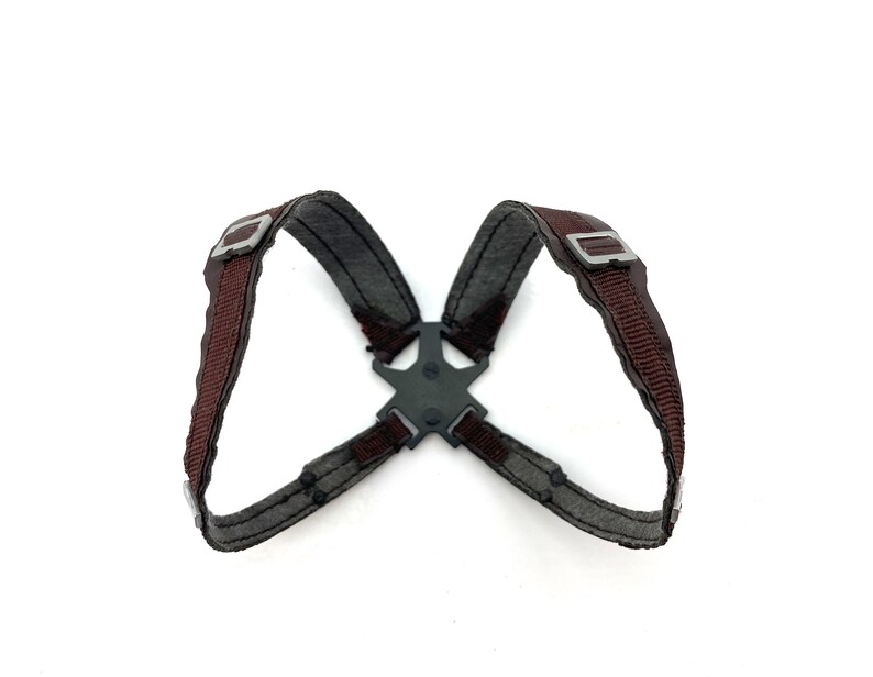 May include: A dark gray and brown harness with metal buckles and a central connector. The harness features brown straps and gray fabric, designed for a secure fit. The design is symmetrical and appears to be made of durable materials.