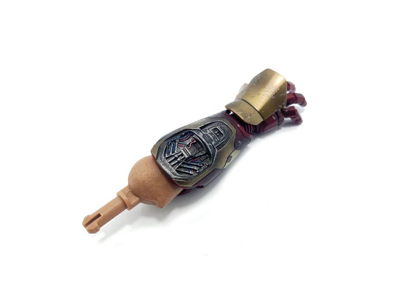 K&ouml;nnte beinhalten: Ein detaillierter Actionfigurenarm mit metallischer Gold- und Burgunder-Farbgebung. Der Arm weist komplizierte mechanische Details und einen hellbraunen Aufsatz auf. Die Hand befindet sich in einer teilweise geschlossenen Position, was auf eine dynamische Pose hindeutet.