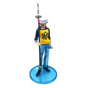 Pu&ograve; includere: Statuetta di un personaggio della serie anime One Piece. La figura indossa una felpa con cappuccio gialla con un emblema nero, jeans blu e un cappello bianco. Tiene in mano un lungo bastone e si trova su una base circolare blu.