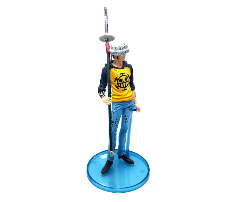 Pu&ograve; includere: Statuetta di un personaggio della serie anime One Piece. La figura indossa una felpa con cappuccio gialla con un emblema nero, jeans blu e un cappello bianco. Tiene in mano un lungo bastone e si trova su una base circolare blu.
