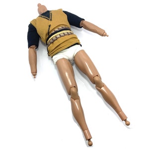 May include: A brown and black action figure with a yellow shirt, black sleeves, and white underwear. The figure has articulated arms and legs.