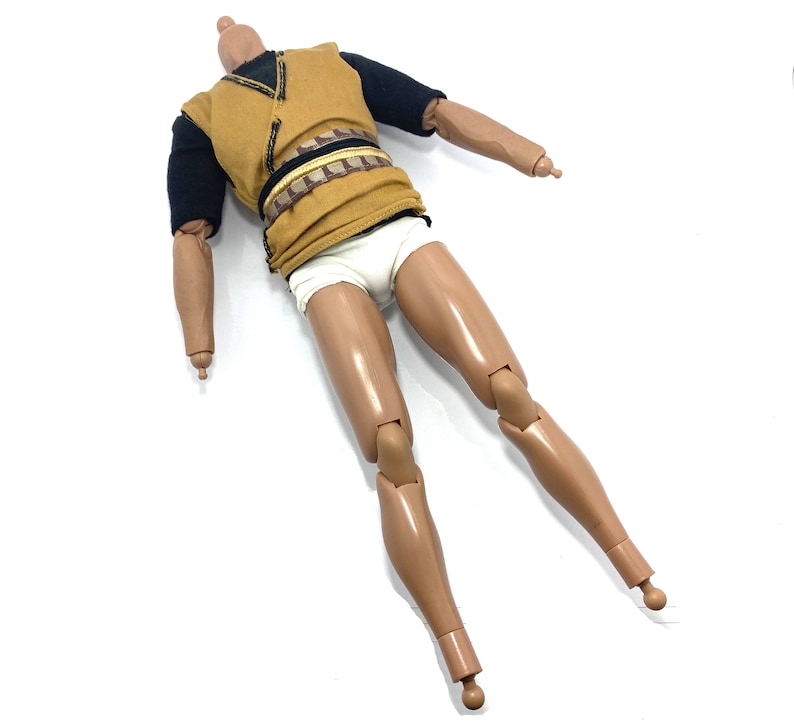 May include: A brown and black action figure with a yellow shirt, black sleeves, and white underwear. The figure has articulated arms and legs.
