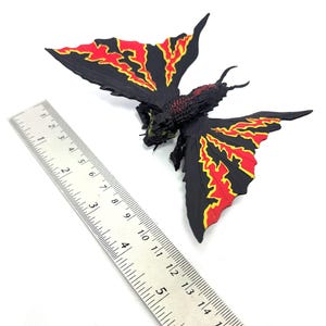 May include: A black moth-like creature figurine with red and yellow flame-patterned wings, positioned next to a metal ruler showing a wingspan of approximately 5 inches. The figure has a textured body, two antennae, and visible legs.