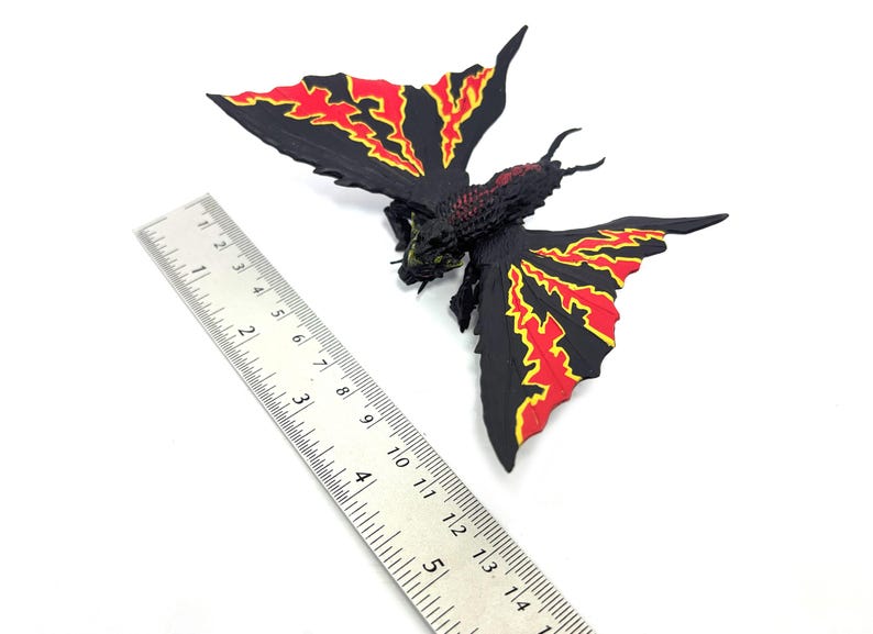 May include: A black moth-like creature figurine with red and yellow flame-patterned wings, positioned next to a metal ruler showing a wingspan of approximately 5 inches. The figure has a textured body, two antennae, and visible legs.