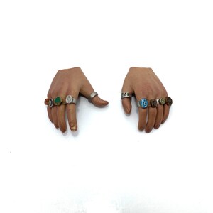 Puede incluir: Dos manos con m&uacute;ltiples anillos en cada dedo. Los anillos son de plata con piedras de diferentes colores, incluyendo verde, azul y marr&oacute;n.