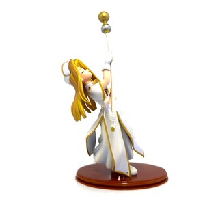 May include: A figurine of a character with long blonde hair, wearing a white and gold outfit, holding a staff with a golden orb. The figurine is on a brown circular base. The character is leaning back, looking up.