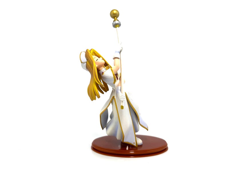 May include: A figurine of a character with long blonde hair, wearing a white and gold outfit, holding a staff with a golden orb. The figurine is on a brown circular base. The character is leaning back, looking up.