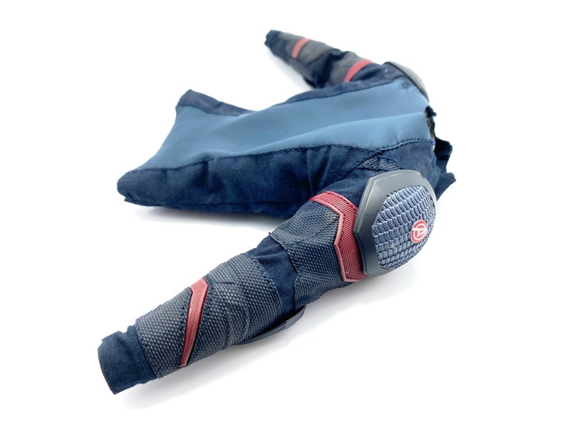 Op de afbeelding: Een zwart en blauw action figure-kostuum met rode accenten en een grijze gevoerde elleboogbeschermer. Het kostuum heeft een textuurstof en een klein rood cirkellogo op de elleboogbeschermer.