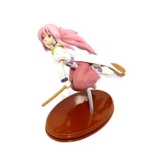 Op de afbeelding: Een beeldje van een vrouwelijk anime-personage met roze haar, gekleed in een witte en groene top, een roze broek en een witte cape. Het figuur staat in een vliegende pose, houdt een houten staf vast en is gemonteerd op een bruine ovale basis.