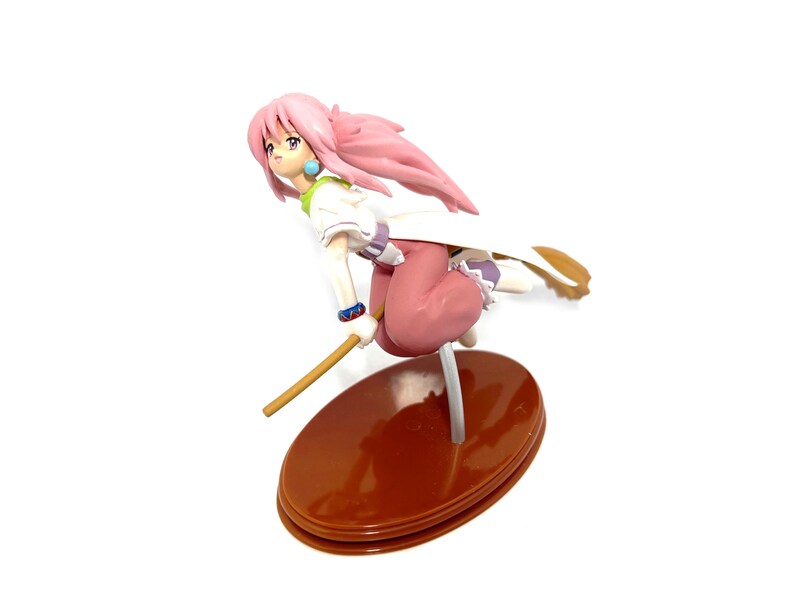 Op de afbeelding: Een beeldje van een vrouwelijk anime-personage met roze haar, gekleed in een witte en groene top, een roze broek en een witte cape. Het figuur staat in een vliegende pose, houdt een houten staf vast en is gemonteerd op een bruine ovale basis.