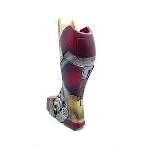 Pu&ograve; includere: Un dettagliato pezzo di armatura per la gamba di Iron Man in rosso metallizzato, oro e argento. L'armatura ha un aspetto usurato con linee di pannelli e rivetti visibili, che suggeriscono un'estetica consumata dalla battaglia. Il design &egrave; molto dettagliato e sembra un oggetto da collezione o da esposizione.