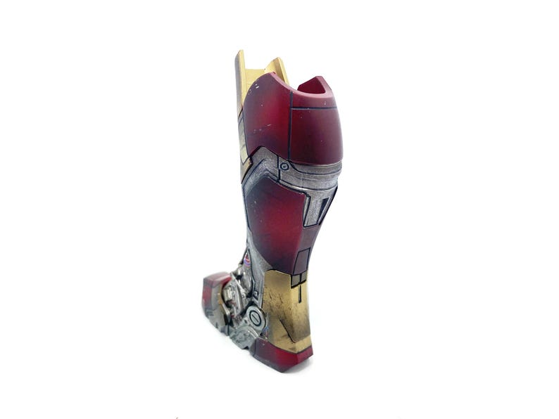 Pu&ograve; includere: Un dettagliato pezzo di armatura per la gamba di Iron Man in rosso metallizzato, oro e argento. L'armatura ha un aspetto usurato con linee di pannelli e rivetti visibili, che suggeriscono un'estetica consumata dalla battaglia. Il design &egrave; molto dettagliato e sembra un oggetto da collezione o da esposizione.
