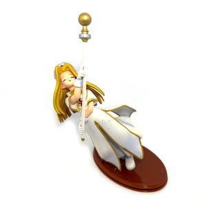 May include: A figurine of a woman in a white dress with gold trim, holding a staff with a gold orb. The figure has long blonde hair and is resting on a brown oval base. The figurine is a collectible item.