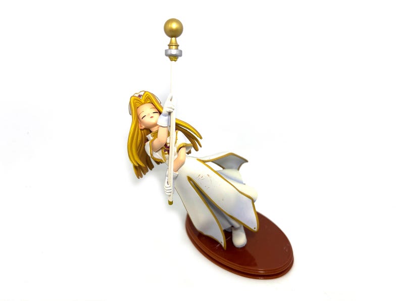 May include: A figurine of a woman in a white dress with gold trim, holding a staff with a gold orb. The figure has long blonde hair and is resting on a brown oval base. The figurine is a collectible item.