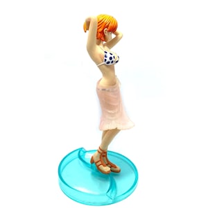 May include: A figurine of a woman with orange hair wearing a pink skirt and a blue and white polka dot bikini top. She is standing on a blue and white base.