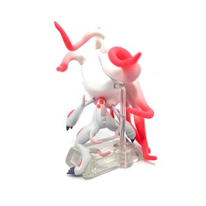 Puede incluir: Una figura blanca y roja de una criatura con un cuerpo esbelto, cuernos curvos y ap&eacute;ndices fluidos en forma de tent&aacute;culos. La figura est&aacute; posada sobre una base rectangular transparente. La paleta de colores incluye blanco, rojo y toques de gris.