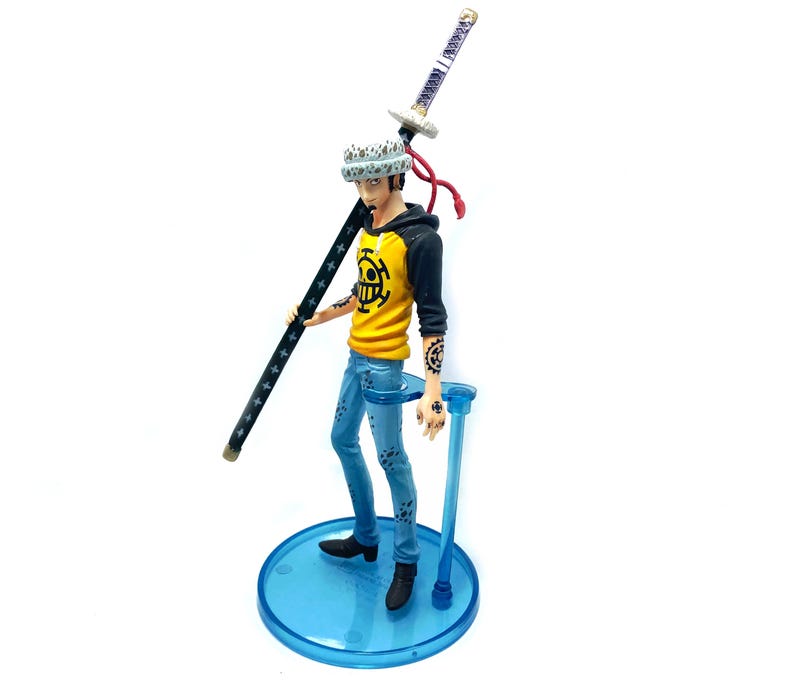 Pu&ograve; includere: Statuetta di un personaggio della serie anime One Piece. La figura indossa una felpa con cappuccio gialla con finiture nere, jeans blu e scarpe nere. La figura tiene in mano un bastone nero con stelle bianche e una spada. La figura &egrave; su un supporto blu.