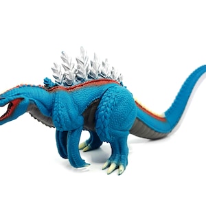 Puede incluir: Un juguete de dinosaurio azul con boca roja y aletas dorsales plateadas. El juguete tiene una cola larga y está posado sobre un fondo blanco. El cuerpo del dinosaurio es texturizado y el juguete probablemente sea de plástico.