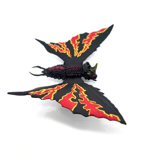 May include: A black insect-like figurine with a textured body and two large wings. The wings feature a prominent red and yellow lightning bolt pattern. The head has a small horn and red eyes, while the tail ends in two curved, pointed pincers.