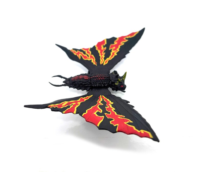 May include: A black insect-like figurine with a textured body and two large wings. The wings feature a prominent red and yellow lightning bolt pattern. The head has a small horn and red eyes, while the tail ends in two curved, pointed pincers.