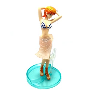 May include: A figurine of a woman with orange hair wearing a blue and white polka dot bikini and a pink sheer skirt. She is standing on a blue and white base.