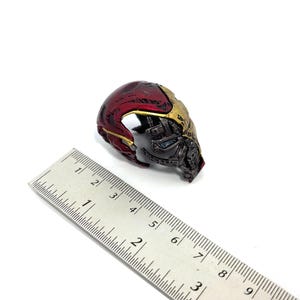 May include: A miniature Iron Man helmet, featuring a metallic red and gold color scheme. The helmet is partially visible, showcasing intricate mechanical details. A ruler is placed next to the helmet for scale.