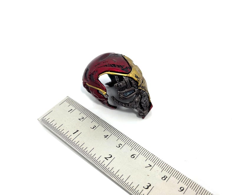 May include: A miniature Iron Man helmet, featuring a metallic red and gold color scheme. The helmet is partially visible, showcasing intricate mechanical details. A ruler is placed next to the helmet for scale.