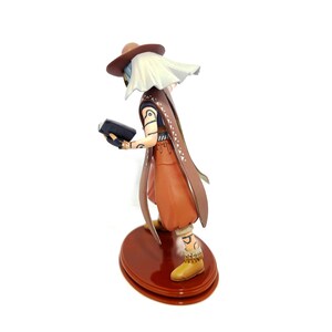May include: A figurine of a character wearing a brown hat, cape, and pants, holding a black book. The figure has white hair and is standing on a brown, circular base. The figure's boots are yellow.