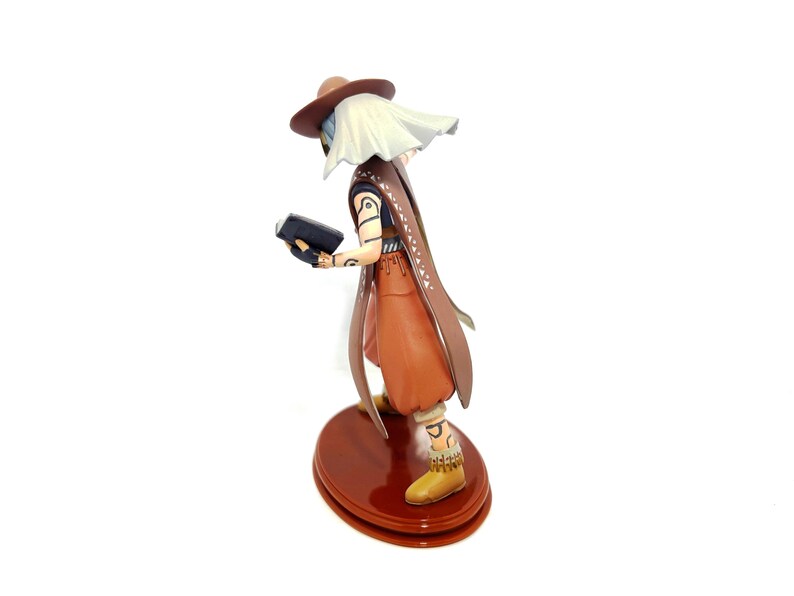 May include: A figurine of a character wearing a brown hat, cape, and pants, holding a black book. The figure has white hair and is standing on a brown, circular base. The figure's boots are yellow.