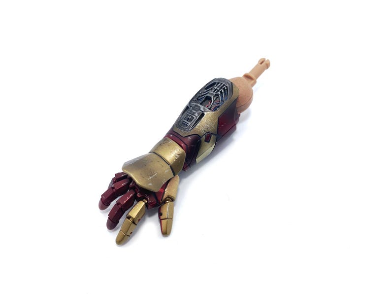 K&ouml;nnte beinhalten: Ein detaillierter Actionfigurenarm mit einer metallischen Gold- und Rotkombination. Der Arm weist komplizierte mechanische Details und eine realistische, bewegliche Hand auf. Die Figur ist vor einem wei&szlig;en Hintergrund platziert.