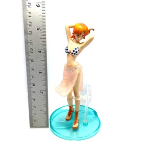 May include: A figurine of a woman with orange hair wearing a pink and white bikini and a pink sarong. She is standing on a blue and white base.
