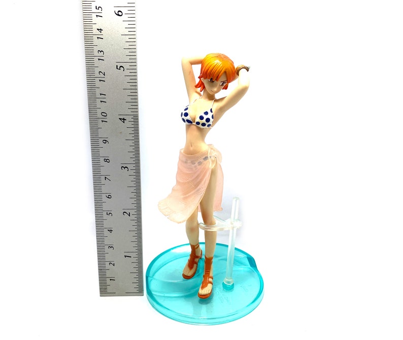 May include: A figurine of a woman with orange hair wearing a pink and white bikini and a pink sarong. She is standing on a blue and white base.