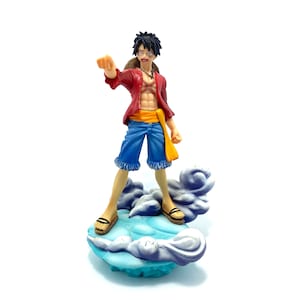 Puede incluir: Una figura de anime roja y azul de un personaje de pie sobre una base de nube azul y blanca.