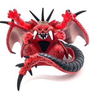 May include: A red monster figurine with black accents, featuring multiple arms, a long tail, and sharp claws. The figurine has a menacing expression with sharp teeth and horns, and small wings. The overall design suggests a fantasy or collectible toy.