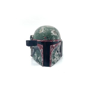 May include: A weathered green and red Boba Fett helmet with a black visor. The helmet has a worn and distressed look, with visible scratches and paint chips.