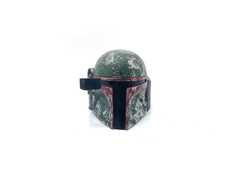 May include: A weathered green and red Boba Fett helmet with a black visor. The helmet has a worn and distressed look, with visible scratches and paint chips.