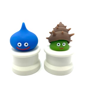 Puede incluir: Dos pequeñas figuritas azules y verdes con caras sonrientes sobre bases blancas. La figurita azul tiene forma de lágrima y la figurita verde tiene forma de caracol.