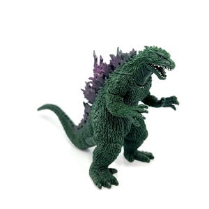 K&ouml;nnte beinhalten: Eine gr&uuml;ne Godzilla-Figur mit einer lila R&uuml;ckenflosse. Das Spielzeug ist in einer bedrohlichen Haltung dargestellt, mit offenem Mund und kleinen wei&szlig;en Z&auml;hnen. Die Figur befindet sich auf wei&szlig;em Hintergrund.