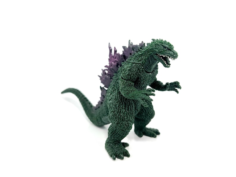 K&ouml;nnte beinhalten: Eine gr&uuml;ne Godzilla-Figur mit einer lila R&uuml;ckenflosse. Das Spielzeug ist in einer bedrohlichen Haltung dargestellt, mit offenem Mund und kleinen wei&szlig;en Z&auml;hnen. Die Figur befindet sich auf wei&szlig;em Hintergrund.