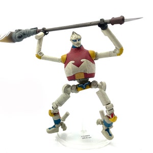 Könnte beinhalten: Eine weiße, rote und gelbe, artikulierte Roboter-Actionfigur, die eine braune und silberne Lanze hält. Der Roboter ist in einer dynamischen Actionpose mit einem erhobenen Bein und der hoch gehaltenen Lanze dargestellt.