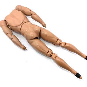 May include: A tan-colored articulated action figure body, lying on a white surface. The figure has a torso, arms, and legs, with joints at the shoulders, elbows, hips, and knees. Black bands are around the ankles. The figure is designed to be posed.