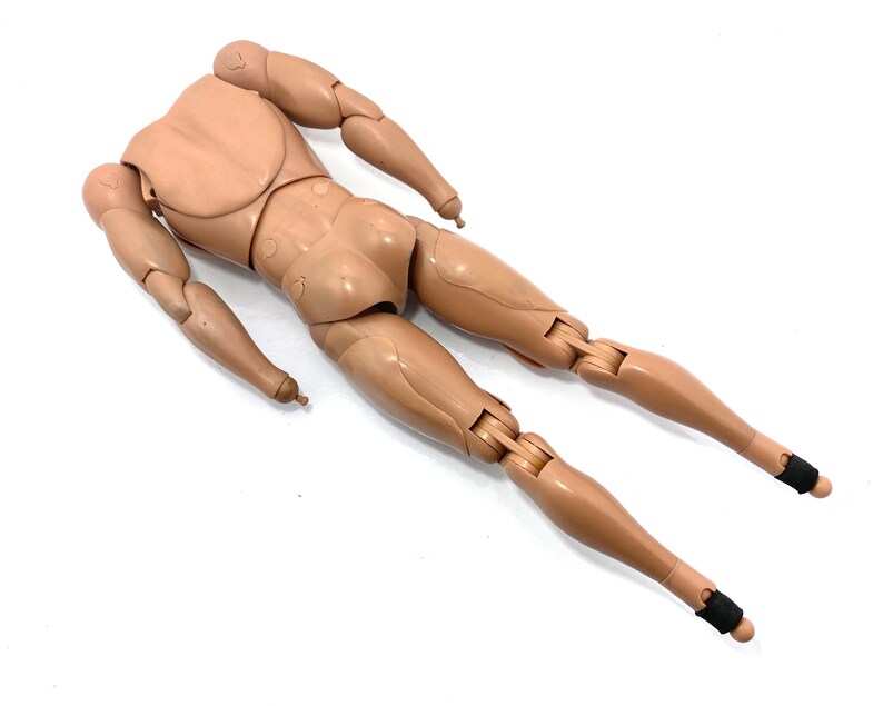 May include: A tan-colored articulated action figure body, lying on a white surface. The figure has a torso, arms, and legs, with joints at the shoulders, elbows, hips, and knees. Black bands are around the ankles. The figure is designed to be posed.