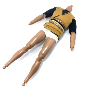 May include: A nude-toned plastic action figure with articulated limbs. The figure is wearing a brown and black tunic with a white undergarment. The figure has a black and brown belt.