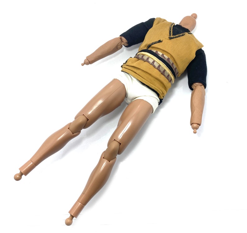 May include: A nude-toned plastic action figure with articulated limbs. The figure is wearing a brown and black tunic with a white undergarment. The figure has a black and brown belt.