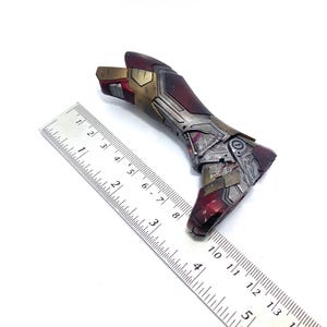 Pu&ograve; includere: Un braccio dettagliato di una action figure metallica nei toni del rosso, oro e argento, con intricati dettagli meccanici. Un righello &egrave; posto accanto al braccio per la scala. Il braccio sembra essere un oggetto da collezione.