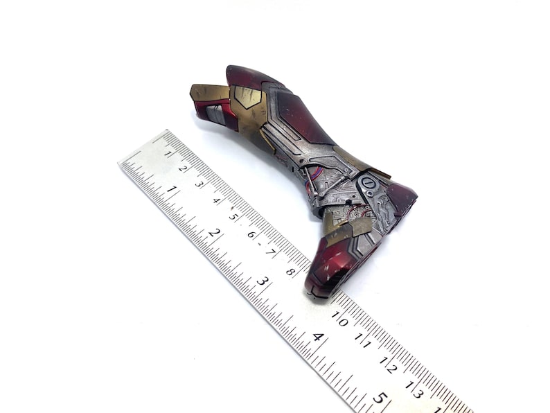 Pu&ograve; includere: Un braccio dettagliato di una action figure metallica nei toni del rosso, oro e argento, con intricati dettagli meccanici. Un righello &egrave; posto accanto al braccio per la scala. Il braccio sembra essere un oggetto da collezione.