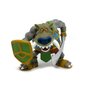 May include: A cartoonish figurine of a troll-like creature, holding a shield and a sword. The figure has a gray body, a green and blue tunic, and yellow sandals. The troll has a large snout, white tusks, and orange eyes.